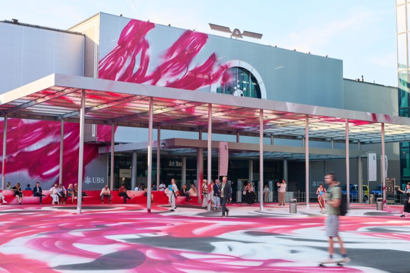 People walk and skateboard outside a modern building in Basel with a large pink and white mural on the walls and ground. The entrance canopy casts shadows, creating a lively and colorful scene that feels like a local hangout. Lifestyle