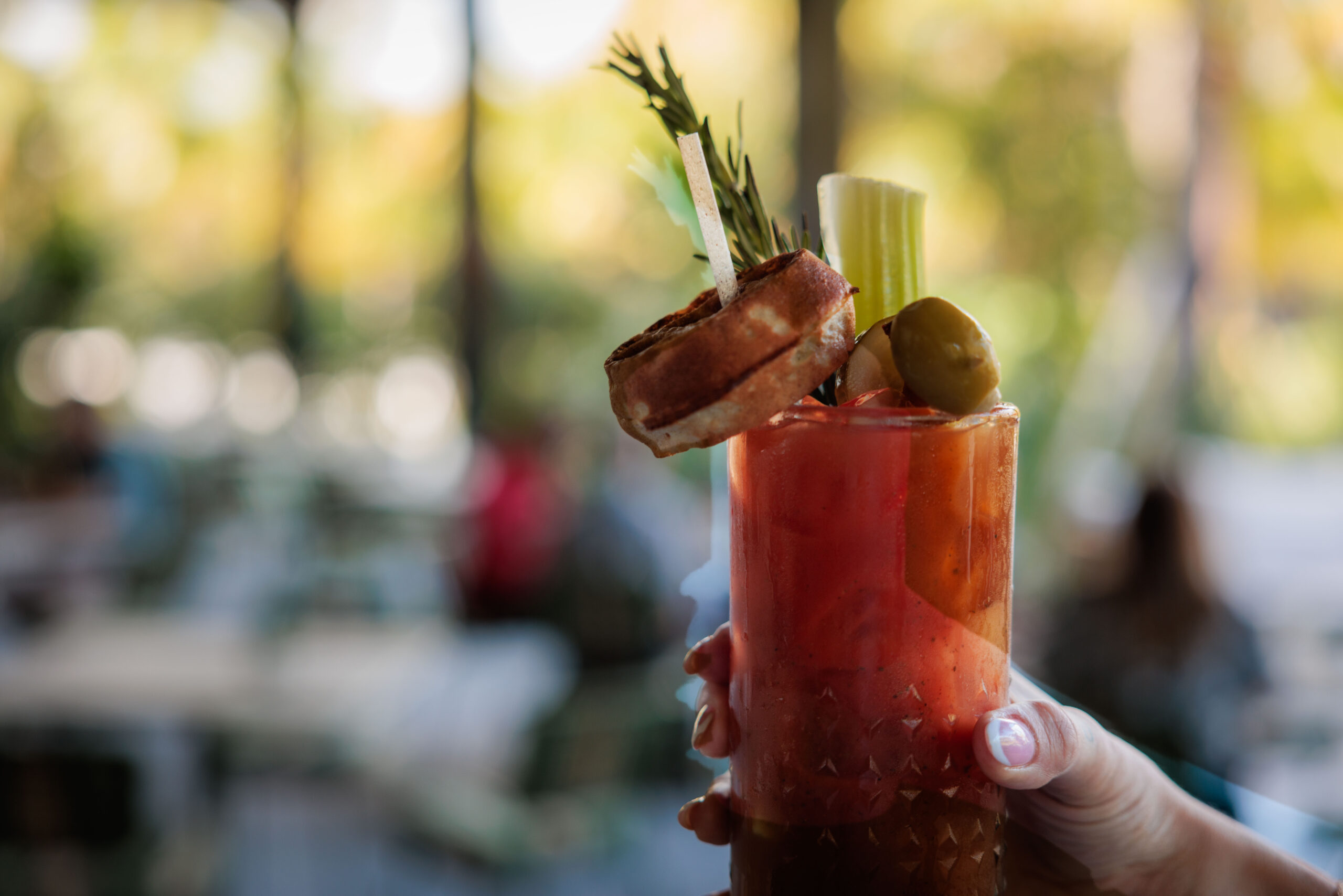 A hand holds a Bloody Mary cocktail garnished with bacon, celery, rosemary, and an olive—an ideal sip for your South Florida holiday break—set against a blurred outdoor background. Lifestyle