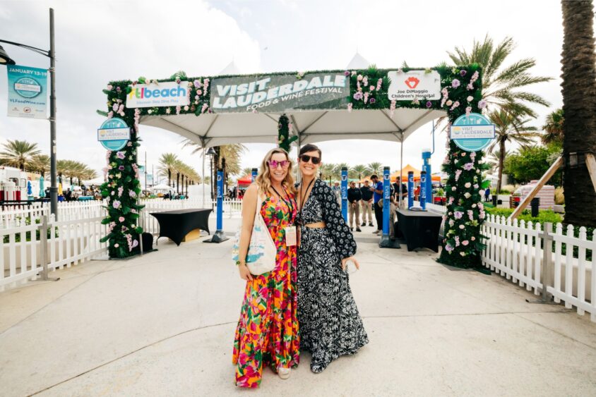 Two smiling women in colorful dresses stand arm in arm under a decorated archway that reads “Visit Lauderdale.” Enjoying the festival under the sun, they’re surrounded by palm trees, white fences, and Taste of Everything signage in the background. Lifestyle