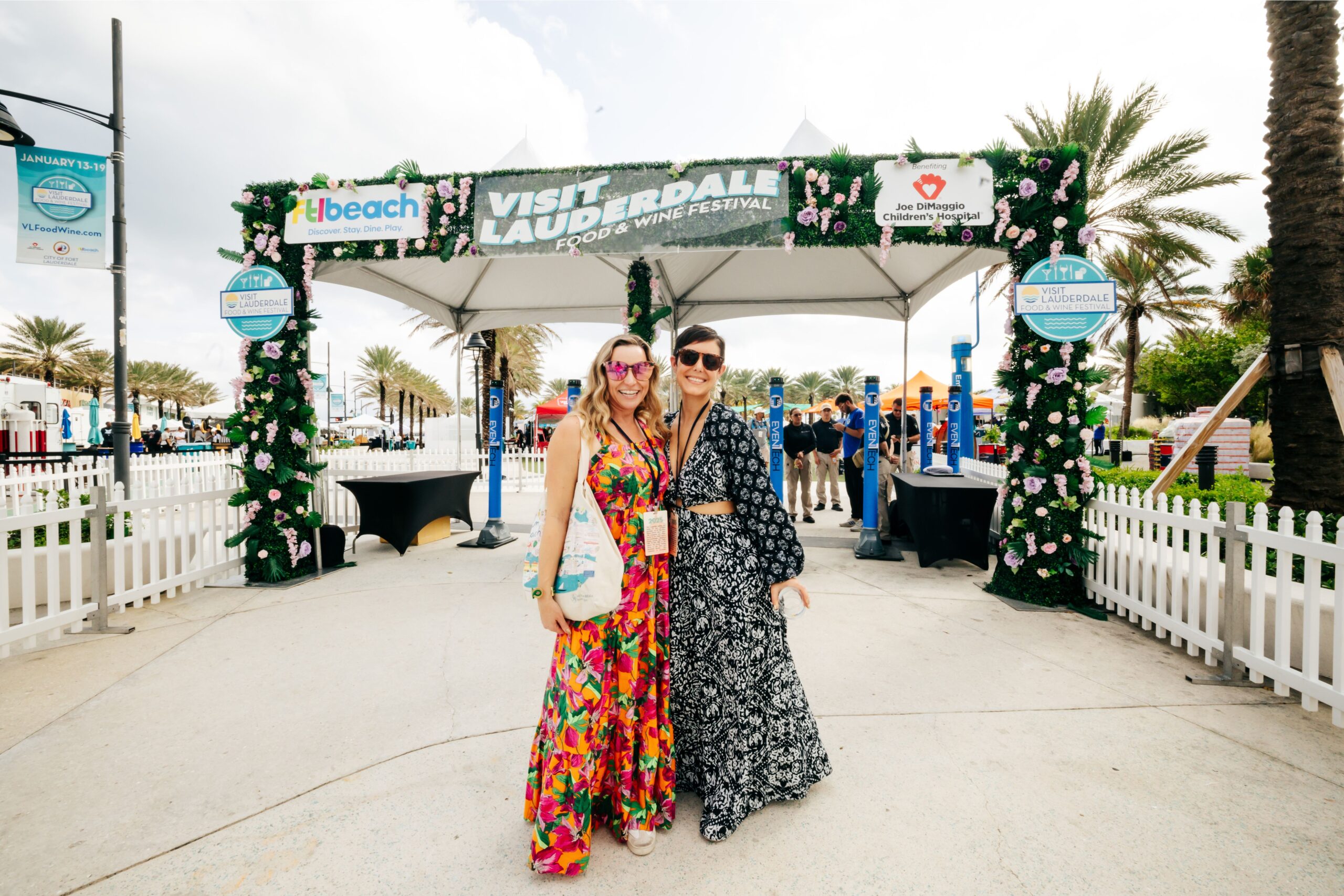 Two smiling women in colorful dresses stand arm in arm under a decorated archway that reads “Visit Lauderdale.” Enjoying the festival under the sun, they’re surrounded by palm trees, white fences, and Taste of Everything signage in the background. Lifestyle