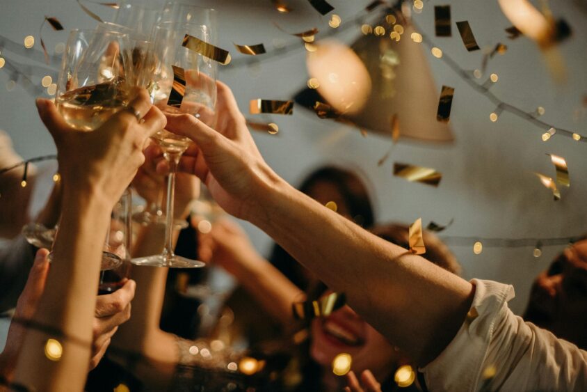 A group of people clink glasses in a celebratory New Year toast, surrounded by golden confetti and string lights, capturing a festive party atmosphere. Lifestyle