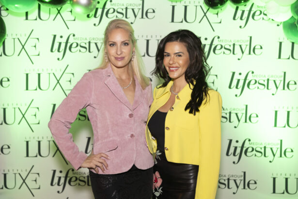Two women pose and smile at a 2025 event in front of a backdrop with "Luxe," "Lifestyle," and Women of Influence logos. One wears a pink blazer, the other a yellow jacket, with green and white balloons visible above. Lifestyle