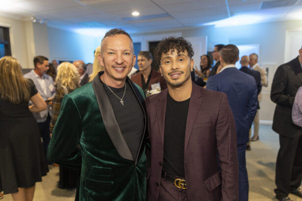 Two men dressed in suits—one in a green velvet jacket and the other in a burgundy suit—smile at the camera during the 2025 Men of Influence indoor event, with people chatting in the background. Lifestyle