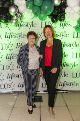 Two women stand in front of a step-and-repeat banner with "LUXE" and "lifestyle" logos, under green and white balloons at the 2025 Women of Influence event. One wears a striped suit, the other a black suit with a red top. Lifestyle