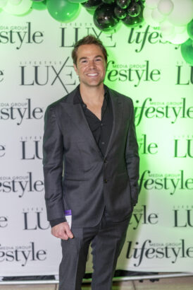 A smiling man in a dark suit stands in front of a backdrop with “LUXE” and “lifestyle” logos and green and white balloons overhead at the 2025 Men of Influence event. Lifestyle