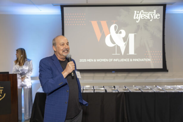 A man in a blue jacket speaks into a microphone at an event, with a screen behind him displaying "2025 Influencers" and "Women of Influence" alongside "lifestyle" magazine branding. A woman stands in the background. Lifestyle