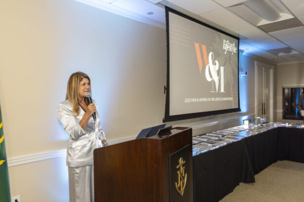 A woman in a silver suit stands at a podium with a microphone, speaking at an event. Behind her, a screen displays “2023 NEW WOMEN OF INFLUENCE HONOREES,” celebrating trailblazers on the path to 2025 Women of Influence. Lifestyle