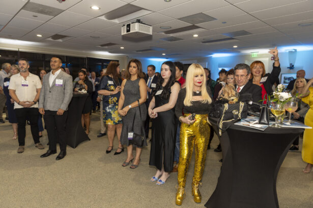 A group of influential people 2025, dressed in business and semi-formal attire, stand in a well-lit event space, watching something off-camera. Some hold drinks near cocktail tables adorned with flowers and name tags. Lifestyle