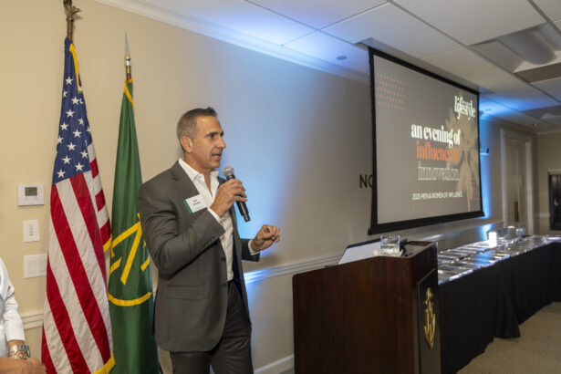 A man in a suit speaks into a microphone at a podium beside American and green flags. Behind him, a screen displays "an evening of influence" at the 2025 Men of Influence indoor event. Lifestyle