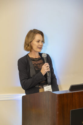 A woman with short, light brown hair stands at a podium, speaking at the 2025 Women of Influence event. She wears a black lace top and blazer with a name tag on her chest, set against a plain, light-colored background. Lifestyle