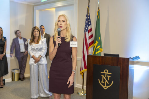 A woman in a sleeveless maroon dress speaks into a microphone at a podium with an anchor logo during the 2025 Women of Influence event, while several people stand in the background near American and green flags. Lifestyle