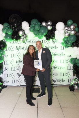 Two people stand smiling on a tiled floor, posing for a photo in front of a green, black, and white balloon arch and a "LUXE lifestyle" backdrop. One person holds a book or magazine at the 2025 Women of Influence event. Lifestyle