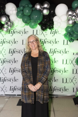 A smiling woman with blonde hair and glasses stands in front of a 2025 "Women of Influence" step-and-repeat banner with "lifestyle" and "LUXE" logos, beneath a green, silver, and black balloon garland. She wears a patterned jacket over a black dress. Lifestyle