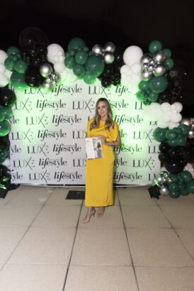 A woman in a yellow dress stands on a tiled floor, holding an open magazine at the 2025 Women of Influence event. Behind her is a "LUXE lifestyle" backdrop and a green, white, black, and silver balloon arch, setting the scene for this elegant gathering. Lifestyle