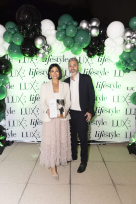 A woman in a light blazer and pink tulle skirt holds a Women of Influence award and stands next to a man in a suit. They pose before a green and white balloon arch and backdrop with "LUXE," "lifestyle," and 2025 event logos. Lifestyle