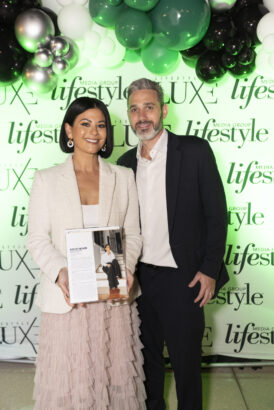 A woman in a light-colored jacket and ruffled skirt holds a magazine while standing next to a man in a dark suit. Smiling, they pose in front of a "Men of Influence 2025" and "LUXE" step-and-repeat backdrop with green and black balloons above. Lifestyle