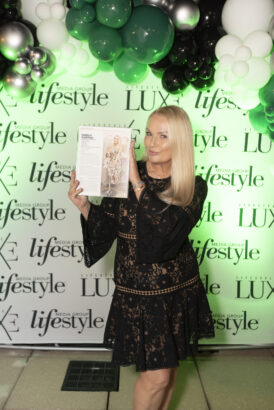 A woman in a black lace dress holds up a magazine featuring herself, standing before a "LUXE" and "lifestyle" backdrop with green, white, and black balloons—celebrating her recognition among the Influential People 2025. Lifestyle