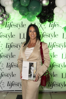 A woman in a tan suit stands holding a magazine and smiling in front of a “Lifestyle Media Group” step-and-repeat backdrop, with white, green, and black balloons above, celebrating the 2025 Women of Influence event. Lifestyle