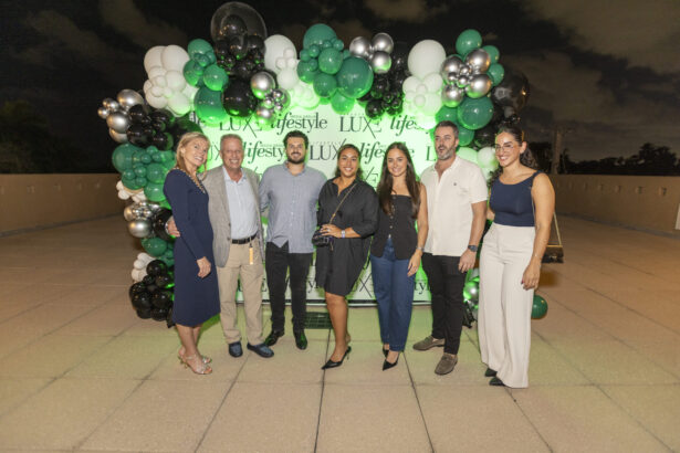 Seven people, including Women of Influence and Men of Influence, pose in front of a green and white balloon arch with a "LUX lifestyle" backdrop at an outdoor nighttime event. They are dressed in semi-formal attire, smiling at the camera, celebrating 2025. Lifestyle
