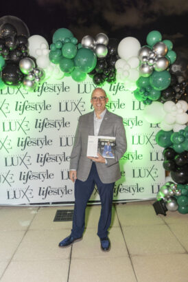 A man in a gray blazer and navy pants stands smiling on a tiled floor, holding a Men of Influence 2025 award, in front of a backdrop with "lifestyle" and "LUXE" logos and green, white, and silver balloon decorations. Lifestyle