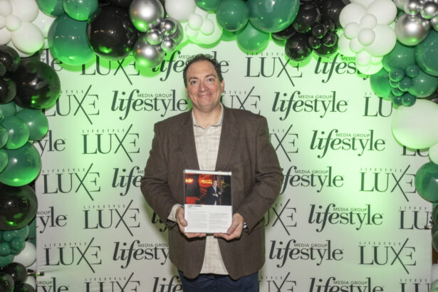 A man in a brown jacket smiles while holding a 2025 Men of Influence magazine at an event, standing in front of a step-and-repeat banner with "LUXE" and "lifestyle" logos, surrounded by green, white, and black balloons. Lifestyle
