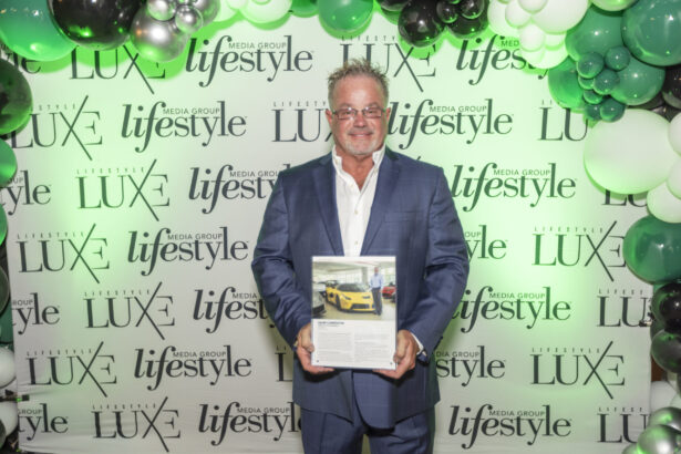 A man in a blue suit stands in front of a “LUXE” and “lifestyle” branded backdrop at the 2025 Men of Influence event, holding a magazine with a yellow sports car on the cover. Green, white, and black balloons decorate the top. Lifestyle