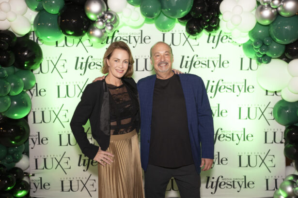 A woman and a man smile at an event, standing in front of a backdrop reading “Lifestyle Luxe,” surrounded by green, black, and white balloon decorations, celebrating the 2025 Women of Influence and Men of Influence honorees. Lifestyle