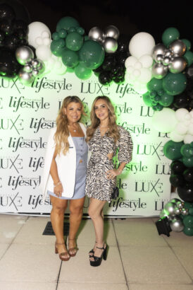 Two women smile and pose together in front of a backdrop with "lifestyle" and "LUXE" logos, surrounded by a decorative arch of black, white, and green balloons at the 2025 Women of Influence event. Lifestyle