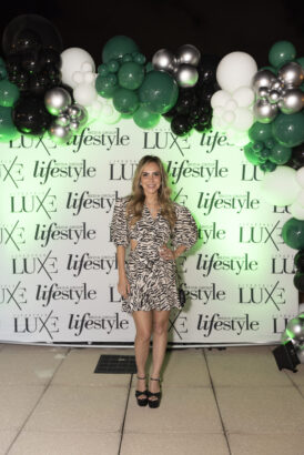 A woman in a patterned dress and black heels poses, smiling, in front of a step-and-repeat banner with “Women of Influence 2025” and “LUXE” logos, decorated with a green, white, and silver balloon arch. Lifestyle