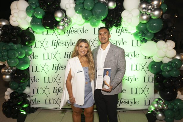 Two people stand smiling in front of a green and white balloon arch and a backdrop featuring "LUXE" and "lifestyle" logos. The man, named a 2025 Men of Influence honoree, holds a magazine open to his photo; the woman wears a white blazer and dress. Lifestyle
