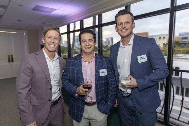 Three men of influence dressed in suits stand together indoors at a social event, smiling at the camera. Two are holding drinks. Large windows behind them reveal a cityscape and water, reflecting the vibrant 2025 networking scene. Lifestyle