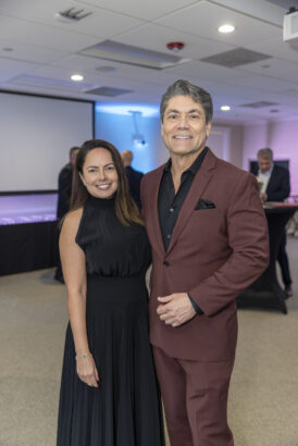 A woman in a sleeveless black dress stands next to a man in a maroon suit with a black shirt. Smiling at the camera, they attend the 2025 Women of Influence event in a well-lit indoor space with other guests in the background. Lifestyle
