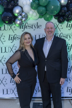 A woman and a man in formal attire, honored as 2025 Women of Influence and Men of Influence, stand smiling before a backdrop with "Lifestyle" and "LUXE" logos, framed by a decorative balloon arrangement above them. Lifestyle