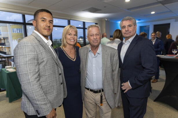 Four people dressed in business attire pose together at an indoor 2025 Men of Influence and Women of Influence networking event, with other attendees and large windows visible in the background. Lifestyle