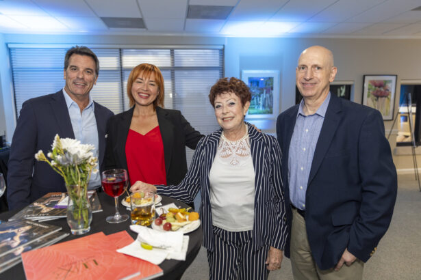 Four adults, three men and one woman, smile and pose together at a table with drinks, fruit, and flowers in a brightly lit indoor setting, capturing a casual gathering of 2025 Men and Women of Influence. Lifestyle