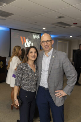 A woman and a man smile while posing together at an indoor Men of Influence and Women of Influence 2025 event. The man wears glasses and a gray blazer; the woman has long brown hair. Other people and a projected presentation screen are visible in the background. Lifestyle