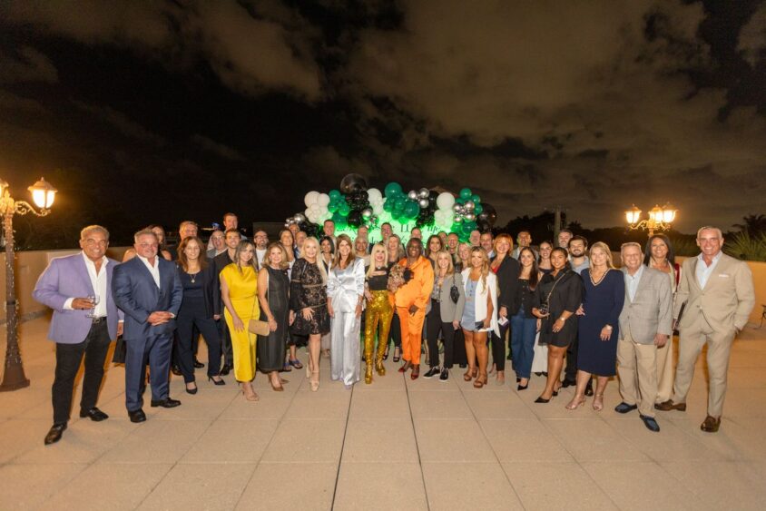 A large group of well-dressed people, including 2025 Men of Influence and Women of Influence, pose outdoors at night on a terrace decorated with white, black, and green balloons under a cloudy sky. Lifestyle