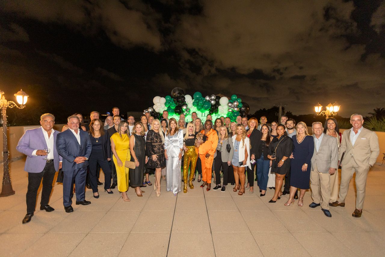 A large group of well-dressed people, including 2025 Men of Influence and Women of Influence, pose outdoors at night on a terrace decorated with white, black, and green balloons under a cloudy sky. Lifestyle