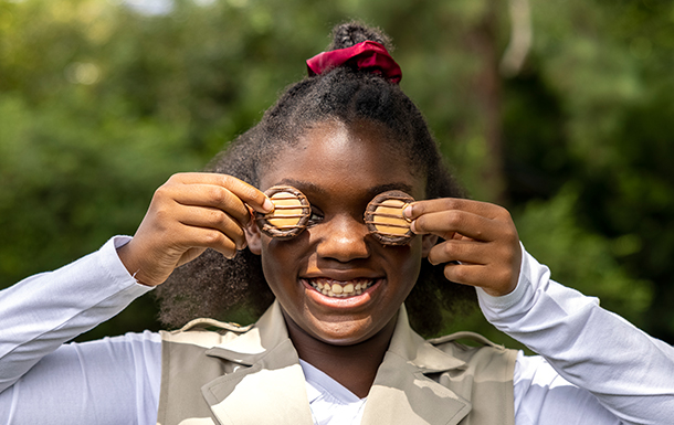 A smiling girl gives a Sweet Salute, holding two chocolate and caramel cookies over her eyes while standing outdoors, with greenery blurred in the background. Lifestyle
