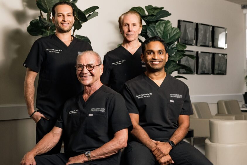 Four men in black medical scrubs smile and pose together in a modern office setting, reflecting modern aesthetics with plants and framed art on the wall behind them. Two are seated in front, while two stand behind. Lifestyle