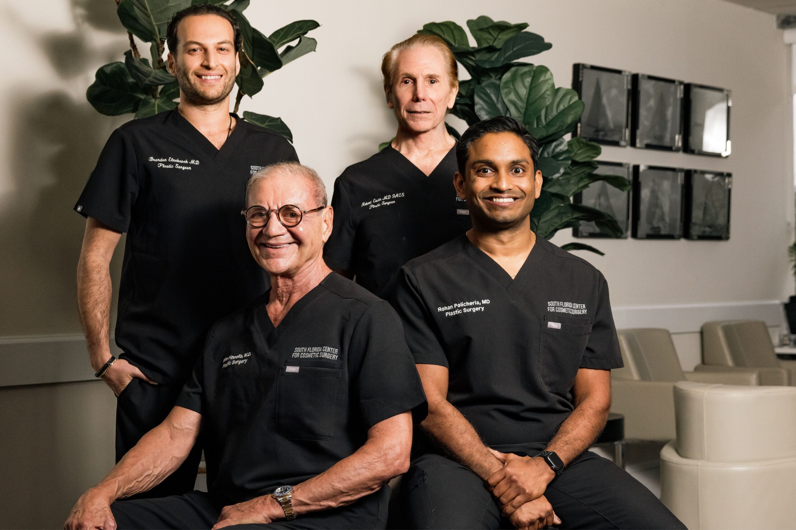 Four men in black medical scrubs smile and pose together in a modern office setting, reflecting modern aesthetics with plants and framed art on the wall behind them. Two are seated in front, while two stand behind. Lifestyle