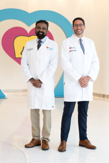Two male doctors, leaders in health care, stand smiling in white lab coats before a wall with a colorful heart design. Both have short hair, wear dress shoes, and pose confidently in a bright, clean hallway. Lifestyle