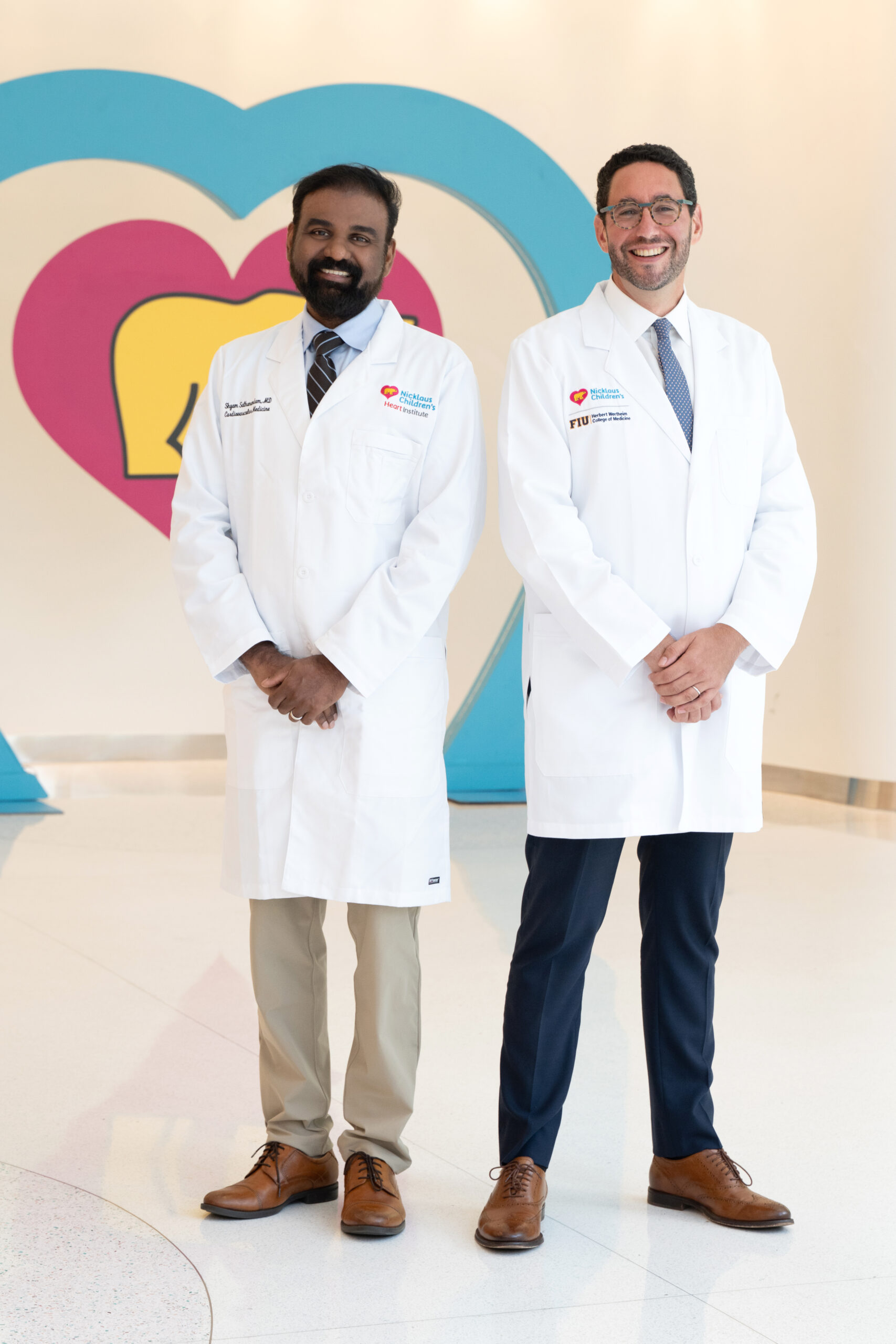 Two male doctors, leaders in health care, stand smiling in white lab coats before a wall with a colorful heart design. Both have short hair, wear dress shoes, and pose confidently in a bright, clean hallway. Lifestyle