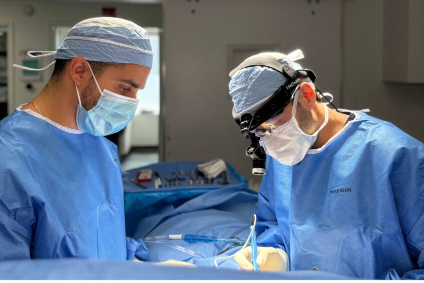 Two surgeons in blue scrubs and masks perform surgery in a well-lit, modern operating room, their focused attention reflecting the art of medicine and modern aesthetics in healthcare. Lifestyle