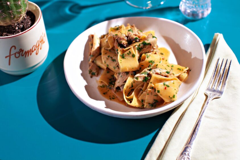 A plate of wide pappardelle pasta with creamy sauce and herbs sits on a blue table beside a fork, pale yellow napkin, glass of water, and a small potted cactus labeled “formaggi”—perfect for celebrating Valentine’s Day Miami style. Lifestyle