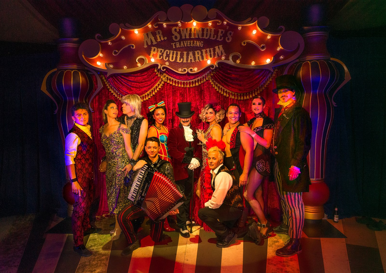 Step right up! A group of performers in colorful, vintage circus costumes pose together under a sign reading “Mr. Swindle’s Traveling Peculiarium,” with bright stage lights and ornate red and gold decorations in the background. Lifestyle