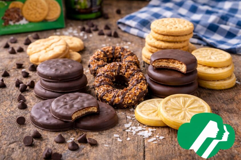 An assortment of Girl Scout Cookies, including chocolate-covered, caramel, and lemon varieties, is arranged on a rustic wooden surface with chocolate chips and a blue plaid cloth. The Girl Scouts logo appears in the corner. Lifestyle