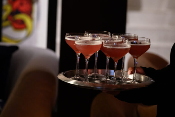 A person holds a silver tray with seven pink cocktails in coupe glasses, each topped with foam, perfect for an After Hours gathering in a dimly lit setting. Lifestyle