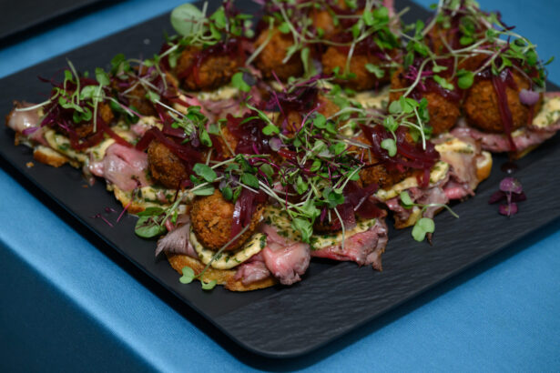 A black platter holds several After Hours appetizers featuring roasted meat, crispy croquettes, microgreens, and a drizzle of sauce, all arranged on small toasted bases atop a blue tablecloth. Lifestyle