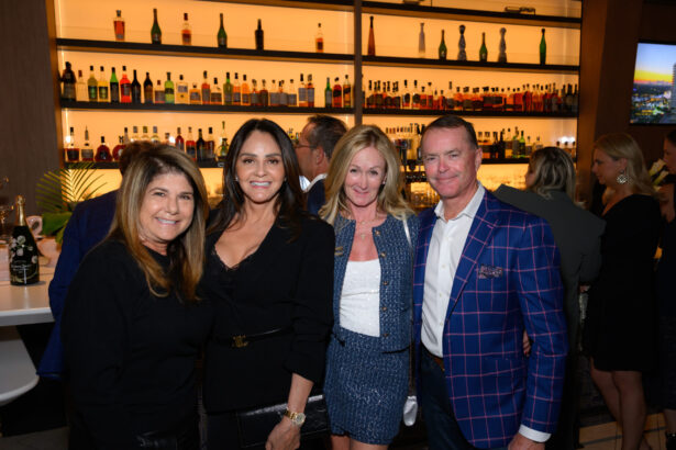 Four people pose and smile together in front of a well-lit bar, capturing the perfect After Hours moment. The two women on the left wear dark outfits, the woman next to them is in blue, and the man on the right sports a blue checked blazer. Lifestyle